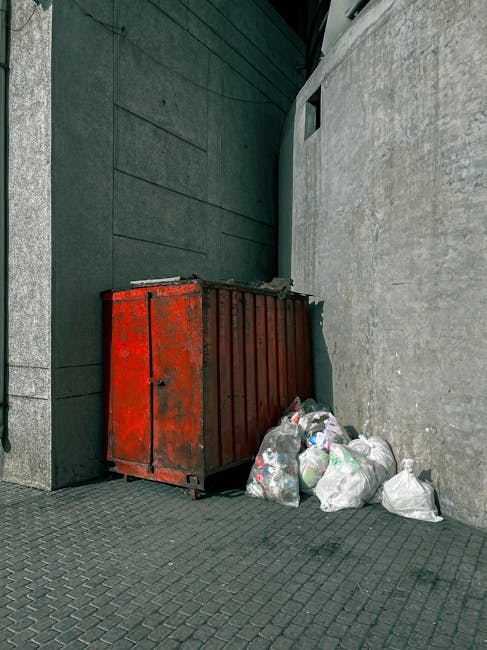 A black refuse wheelie bin with the label 'ST. JOHN'S' positioned on a residential pavement at night, filled with various discarded household items and rubbish bags. The bin's surface appears slightly