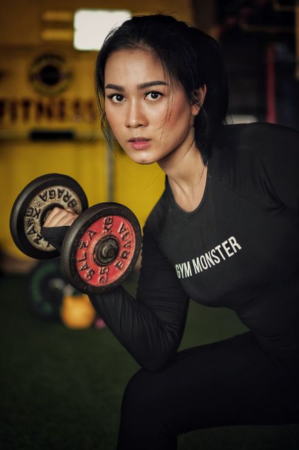A young woman with dark hair tied back, wearing a black long-sleeved shirt with the words 'GYM MONSTER' printed on it, is performing a bicep curl with a black and red dumbbell. She is indoors in a gym