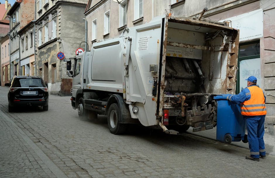 A person wearing orange work overalls and white shoes is standing on a grey concrete floor, holding two large blue plastic rubbish bags, one in each hand, which are filled with waste and appear slight